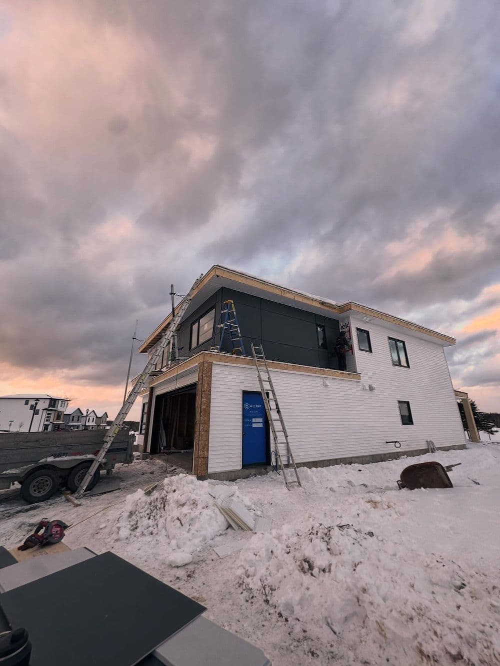 Construction site of a modern home with ladders, workers, and snowy surroundings at sunset.