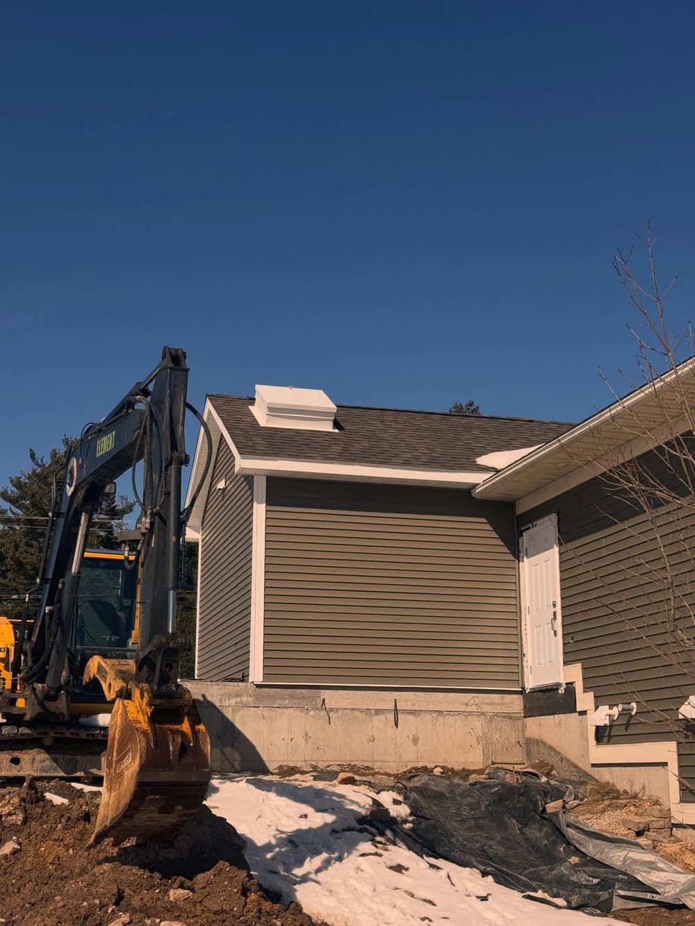 Excavator near a home under renovation, blue sky, and snow on the ground.