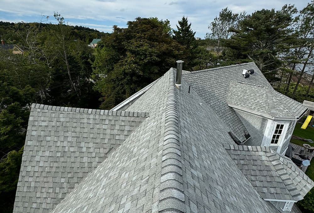Aerial view of a gray shingle roof with trees and a chimney in the background.