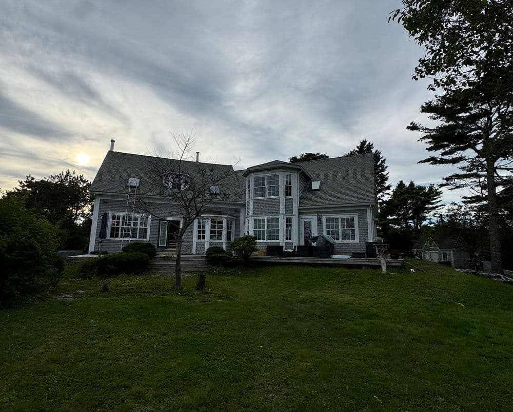 Elegant gray house with large windows surrounded by green lawn and trees under a cloudy sky.